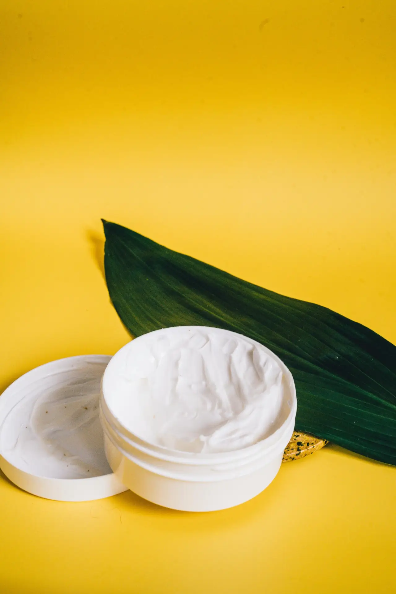 Open cosmetic cream container with a green leaf on a vibrant yellow backdrop, showcasing natural beauty.